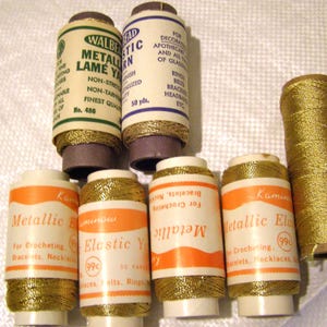 May include: A collection of seven spools of metallic gold thread and elastic yarn. Two spools feature white labels with green and blue text, while four spools have orange and white labels with black text. One spool is wrapped in gold thread without a label.