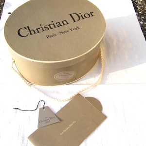 May include: A tan round box with the words "Christian Dior Paris - New York" printed in black. A tag and a card are next to the box. The box has a cream-colored rope handle.