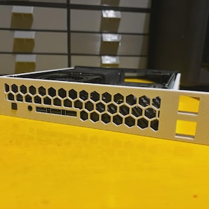 Może przedstawiać: Srebrna obudowa komputera z wzorem plastra miodu na przednim panelu. Obudowa posiada kilka portów i otworów i jest umieszczona na żółtej powierzchni. Została zaprojektowana do umieszczania komponentów komputera.