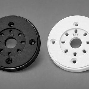 May include: Two circular components, one black and one white, each with multiple holes and the letters "TZE" in the center. The black component has a matte finish, while the white one appears smooth. Both are set against a gray background.