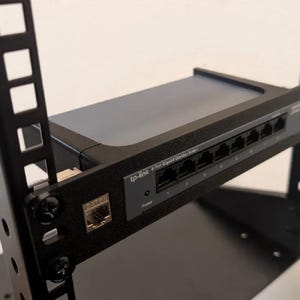 May include: A black network rack with a grey and black TP-Link 8-Port Gigabit Ethernet Switch. The switch is mounted on a shelf within the rack. The rack has a series of square and round holes along the sides.
