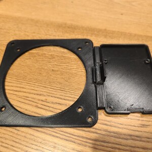 May include: Black plastic frame, rectangular with a circular aperture and a hinged rectangular section. The frame has several small holes for mounting. Likely a component or part of a larger device.