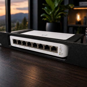 May include: A black and white network switch with multiple ports, housed in a black rack mount. The device has indicator lights and is set on a dark wooden surface. A black mug and a camera lens are also visible.