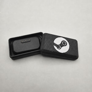 May include: Black rectangular case with a hinged lid, open to reveal a smaller black compartment. The lid features a white Steam logo. The case is likely for storing or protecting a small electronic device.