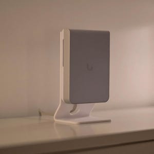 May include: A white and gray rectangular device with a rounded top, mounted on a white stand. The device has a small, circular logo on the front. The stand has a curved design and is placed on a white surface.