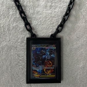 May include: Black necklace featuring a rectangular pendant holding a trading card. The card displays a colorful illustration of a dragon-like creature with the text "Mega Charizard X". The chain is made of black, chunky links.