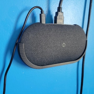 May include: A black oval-shaped electronic device with a fabric-covered front and a small circular logo in the center. The device is mounted in a black plastic holder and is connected to two black cables, one of which is labeled HDMI at the connector.