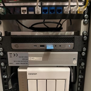 May include: A server rack with various electronic components. Visible are network cables, an HP Elitedesk, a silver device with a label, and a white QNAP storage device. The rack is mounted with screws.