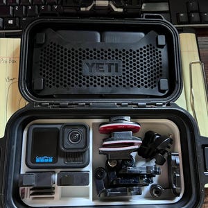 May include: Black YETI Pro Box open, revealing a GoPro camera and various accessories. The camera is black with a blue GoPro logo. Accessories include mounts and a red and white component. The box is on a desk.