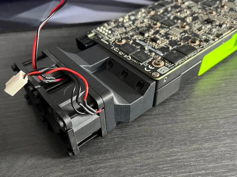 Op de afbeelding: Een zwarte en grijze computer grafische kaart met een groene accentkleur. De kaart heeft een koelventilator en is verbonden met draden met een witte connector. De componenten liggen op een donkere ondergrond.
