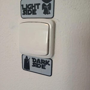 Puede incluir: Un interruptor de luz blanco con dos pegatinas rectangulares arriba y abajo. La pegatina superior dice "LIGHT SIDE" con una silueta de Yoda, y la pegatina inferior dice "DARK SIDE" con una silueta de Darth Vader. Las pegatinas son grises con texto e im&aacute;genes en negro.