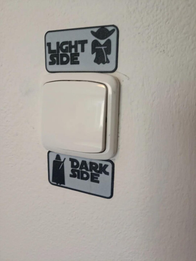 Puede incluir: Un interruptor de luz blanco con dos pegatinas rectangulares arriba y abajo. La pegatina superior dice "LIGHT SIDE" con una silueta de Yoda, y la pegatina inferior dice "DARK SIDE" con una silueta de Darth Vader. Las pegatinas son grises con texto e im&aacute;genes en negro.