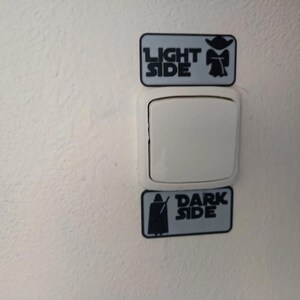 Puede incluir: Un interruptor de luz blanco con dos pegatinas rectangulares arriba y abajo. La pegatina superior dice "LIGHT SIDE" con un gr&aacute;fico de Yoda. La pegatina inferior dice "DARK SIDE" con un gr&aacute;fico de Darth Vader. Las pegatinas son de color azul claro con texto y gr&aacute;ficos negros.
