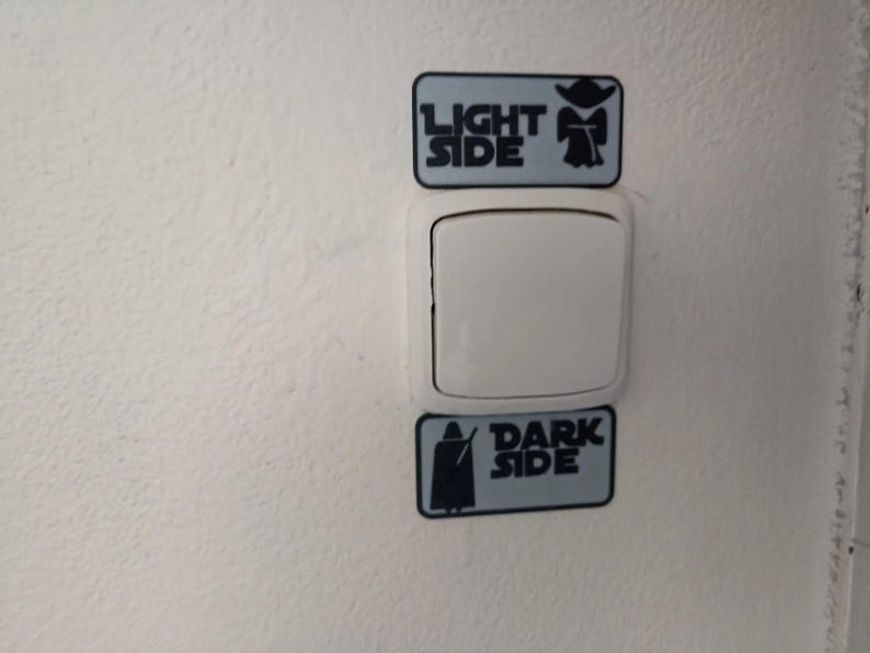 Puede incluir: Un interruptor de luz blanco con dos pegatinas rectangulares arriba y abajo. La pegatina superior dice "LIGHT SIDE" con un gr&aacute;fico de Yoda. La pegatina inferior dice "DARK SIDE" con un gr&aacute;fico de Darth Vader. Las pegatinas son de color azul claro con texto y gr&aacute;ficos negros.