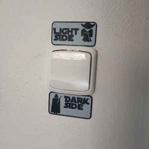 Puede incluir: Un interruptor de luz blanco con dos pegatinas rectangulares arriba y abajo. La pegatina superior dice "LIGHT SIDE" con una silueta de Yoda. La pegatina inferior dice "DARK SIDE" con una silueta de Darth Vader. Las pegatinas son grises y negras.