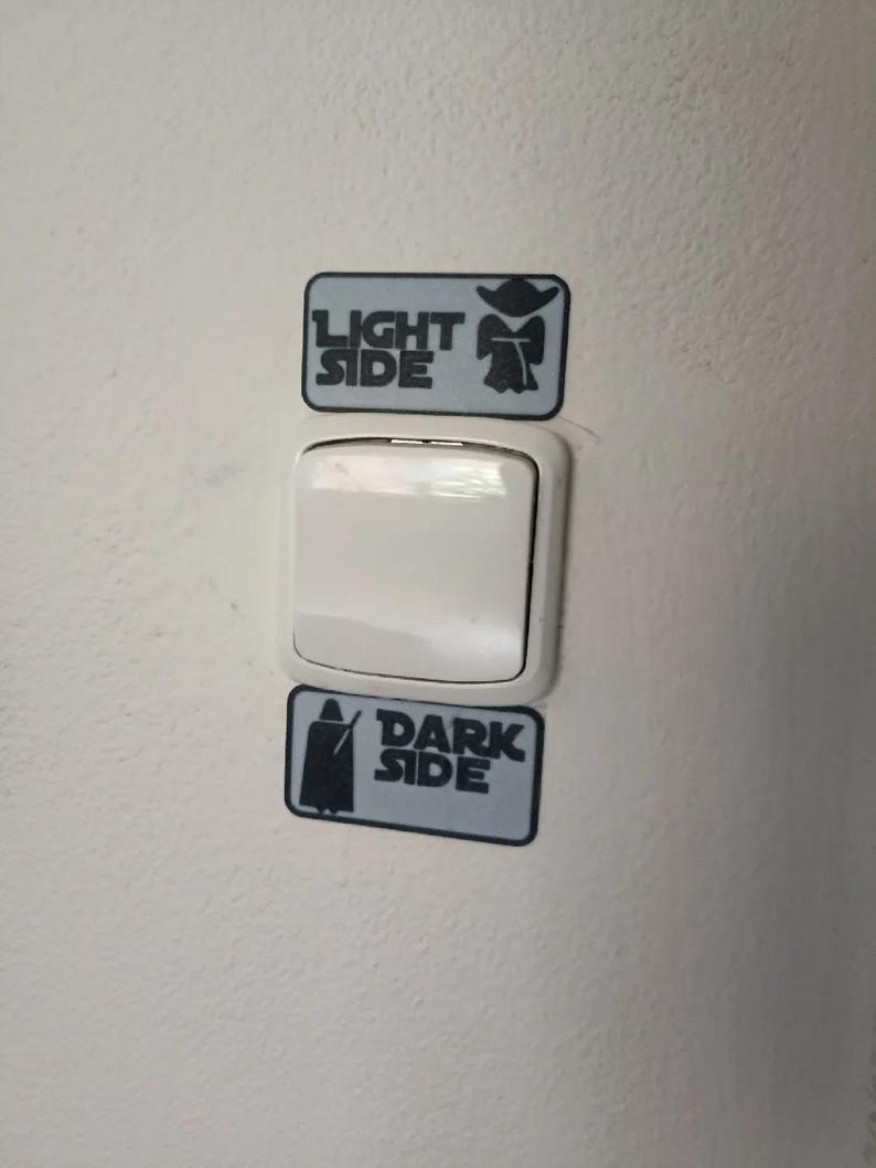 Puede incluir: Un interruptor de luz blanco con dos pegatinas rectangulares arriba y abajo. La pegatina superior dice "LIGHT SIDE" con una silueta de Yoda. La pegatina inferior dice "DARK SIDE" con una silueta de Darth Vader. Las pegatinas son grises y negras.