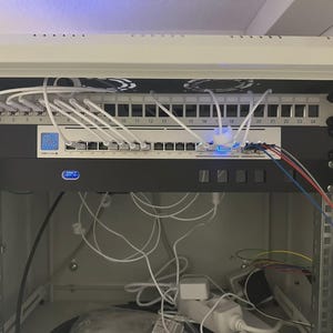 May include: A network rack with multiple network switches, patch panels, and a variety of cables. White Ethernet cables are connected to the switches. The rack is mounted inside a white cabinet. The image shows a complex network setup.