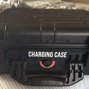May include: A black rectangular hard-shell protective case with a reinforced grid pattern on the lid. The front features a black label with the white text CHARGING CASE and a circular pressure release valve with a red ring and small text.