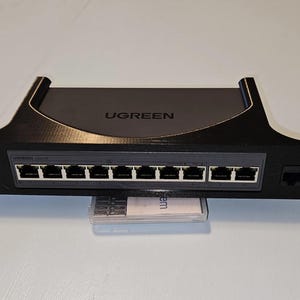 May include: A black UGREEN network switch mount featuring a central panel with eight Ethernet ports and two additional side ports. The mount has a curved top edge and mounting holes on each side, designed to hold networking hardware securely.