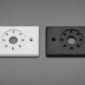 May include: Two rectangular electrical outlet covers, one white and one black, are displayed side-by-side. Each cover has a circular pattern of holes in the center and two smaller holes on either side. The covers are made of a solid material.
