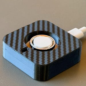 May include: A square-shaped charging dock featuring a black and blue carbon fiber pattern on the top surface. The device has a central circular cutout containing a white button and is connected to a white power cable extending from the side.