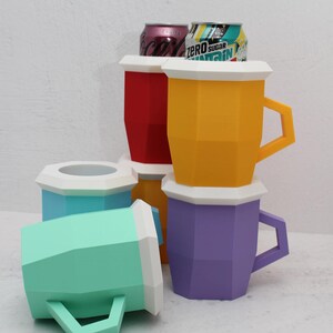 May include: A collection of geometric, faceted beverage holders in red, yellow, purple, light blue, and mint green, each featuring a white rim and angular handle. Two aluminium soda cans are visible inside the red and yellow holders.