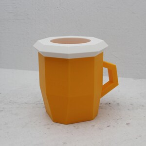 May include: A geometric mug with a faceted orange body and a white lid featuring a circular opening. The mug has a sharp, angular handle and a distinct polygonal shape, presenting a modern, structured design with clean lines and a solid colour finish.