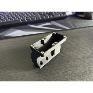 Op de afbeelding: Een donkergrijs rechthoekig object met afgeronde hoeken en verschillende kleine, ronde gaten. Het object heeft goudkleurige accenten en lijkt een onderdeel of behuizing te zijn voor een groter apparaat. Een toetsenbord en muis zijn zichtbaar op de achtergrond.