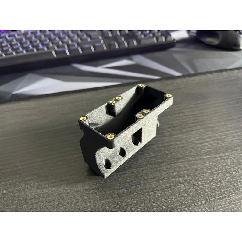 Op de afbeelding: Een donkergrijs rechthoekig object met afgeronde hoeken en verschillende kleine, ronde gaten. Het object heeft goudkleurige accenten en lijkt een onderdeel of behuizing te zijn voor een groter apparaat. Een toetsenbord en muis zijn zichtbaar op de achtergrond.