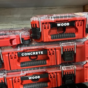May include: Several red toolboxes with clear lids are stacked. The toolboxes have black latches and handles. The labels on the toolboxes read "WOOD" and "CONCRETE". The toolboxes are on a shelf.