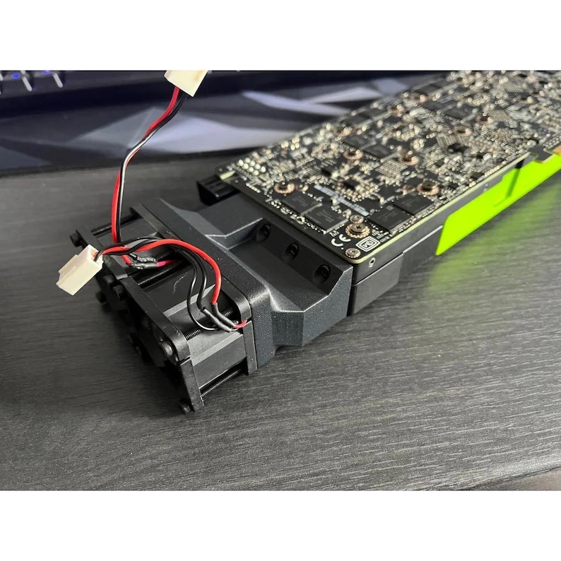 Op de afbeelding: Een zwarte en groene computer grafische kaart met een zwarte ventilator en blootliggende bedrading. De kaart heeft een complexe printplaat met talrijke kleine componenten. De ventilator is bevestigd aan de kaart met rode en zwarte draden en witte connectoren.
