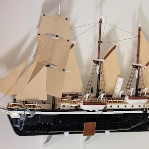 Könnte beinhalten: Detailliertes Modell eines Großseglers mit beigen Segeln, schwarzem Rumpf und weißen Akzenten. Das Schiff verfügt über mehrere Masten, Takelage und eine rote Flagge. Das Modell ist an der Wand befestigt.