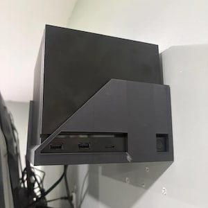 Op de afbeelding: Een matzwarte wandhouder voor consoles met een geometrisch, hoekig ontwerp. De beugel houdt een rechthoekig elektronisch apparaat stevig vast, waarbij twee USB-poorten aan de voorzijde en een kleine horizontale gleuf in het voorpaneel zichtbaar zijn.