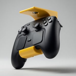 Pode incluir: Um controle de videogame preto é montado em um suporte amarelo. O controle possui vários botões, joysticks e um direcional. O suporte foi projetado para segurar o controle com segurança.