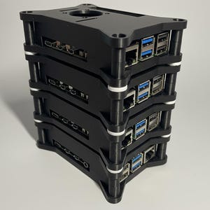 May include: A black, stacked electronic device with multiple ports and slots. The device has a rectangular shape with a series of layers held together by vertical supports. White accents are visible between the layers. The device is likely a computer component or server.