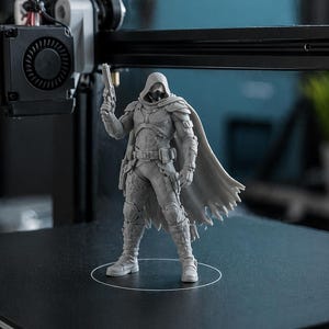 May include: A grey 3D-printed figurine of a character in a hooded cloak, holding a gun. The figure is standing on a black surface, with a 3D printer in the background. The figurine has detailed armour and a flowing cape.