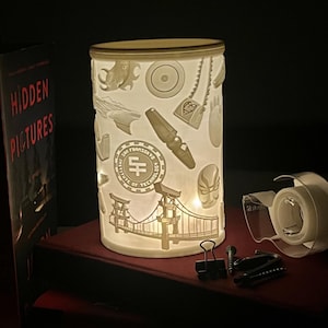 May include: A cylindrical lamp with a cream-colored shade featuring etched designs of various objects, including a pen, a bridge, and a logo. The lamp is lit from within, casting a warm glow. The lamp is sitting on top of a stack of books.