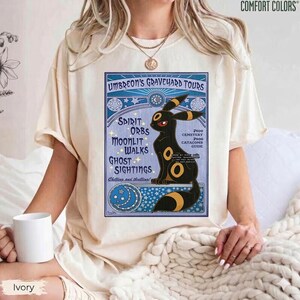 May include: Ivory-colored t-shirt featuring a graphic of Umbreon's Graveyard Tours. The design includes a black Umbreon character with yellow accents, set against a blue background with text that reads "Spirit Orbs, Moonlit Walks, Ghost Sightings." The shirt is made by Comfort Colors.