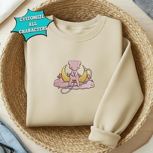 May include: Beige sweatshirt with an embroidered design featuring a pink creature sitting on a crescent moon and cloud. The design includes gold and purple accents. The text "CUSTOMIZE ALL CHARACTERS" is in a blue starburst.
