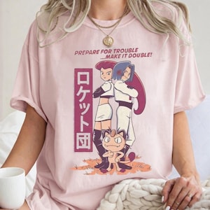 May include: A light pink t-shirt featuring a cartoon graphic of two characters and a Meowth. The text "PREPARE FOR TROUBLE... MAKE IT DOUBLE!" is above the characters. Japanese text is also present.