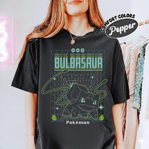May include: Black t-shirt with a green and blue Bulbasaur graphic and the word "Bulbasaur" in a retro font. The word "Pokémon" is printed below the graphic. The shirt is from the Comfort Colors Pepper collection.