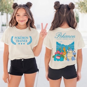 Puede incluir: Camiseta color crema con la inscripción "Pokémon Trainer" en azul, enmarcada por una corona de laurel. La parte trasera de la camiseta muestra la palabra "Pokémon" sobre un gráfico de varios personajes Pokémon.
