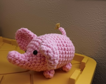 Könnte beinhalten: Ein hellrosa gehäkeltes Stofftier in Form eines Elefanten mit großen Ohren, einem Rüssel und einem kleinen gelben Garnbüschel am Ende des Schwanzes. Der Elefant hat zwei kleine schwarze Kunststoffaugen und eine strukturierte, geknotete Oberfläche.
