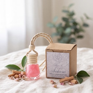 Op de afbeelding: Een klein glazen flesje gevuld met roze vloeistof, voorzien van een houten dop en een ophangkoord. Daarnaast staat een bruine kartonnen doos met een etiket met de tekst Fleur de Sakura, Diffuseur suspendu parfumé, Parfum de Grasse sans CMR, sans phtalates et cruelty free.