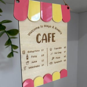 May include: Wooden cafe sign with a pink and yellow awning design. The sign reads "Welcome to Maya & Rivah's CAFE" and lists menu items like Babychino, Fizzy, Juice, and Milkshake. The sign is suspended by a brown rope.