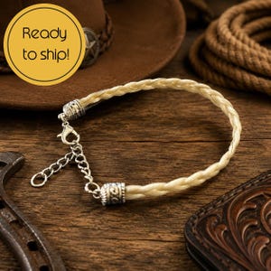 May include: A braided cream-colored bracelet featuring silver-toned metal end caps and a lobster claw clasp with an adjustable chain link extension. The bracelet is circular in shape and displays a textured, woven design throughout the band.