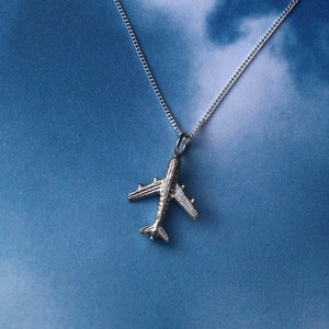 May include: A silver airplane necklace on a silver chain against a blue and white cloudy background.