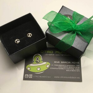 May include: A pair of silver skull earrings in a black box with a green ribbon. The box has the text "OTRO MUNDO" and "JOYERIA CONTEMPORANEA" on it. The box is sitting on a white surface.