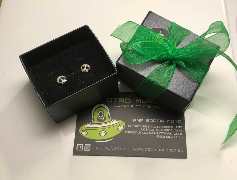May include: A pair of silver skull earrings in a black box with a green ribbon. The box has the text "OTRO MUNDO" and "JOYERIA CONTEMPORANEA" on it. The box is sitting on a white surface.