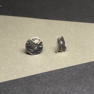 May include: A pair of silver earrings, one shaped like a rocket and the other a textured circle.